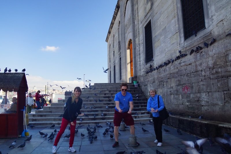 That Backpacker Audrey Bergner, Nomadic Samuel, and Samuel’s mom feeding pigeons on the steps of a historic stone mosque in Istanbul, Turkey, enjoying a playful travel moment surrounded by fluttering birds