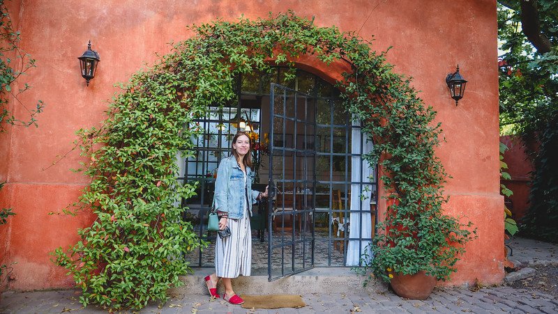Audrey Bergner of That Backpacker standing at the arched entrance of a terracotta building covered in vines at Finca Adalgisa in Mendoza, Argentina. She’s holding the door open, wearing a denim jacket and striped dress while exploring the hotel grounds