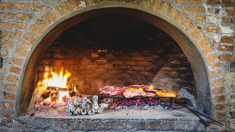 An Argentine asado cooking inside a traditional brick fireplace at Finca Adalgisa in Mendoza, Argentina. Cuts of meat, lemons, and foil-wrapped vegetables are grilled over hot embers, capturing the essence of Argentina’s famous barbecue tradition