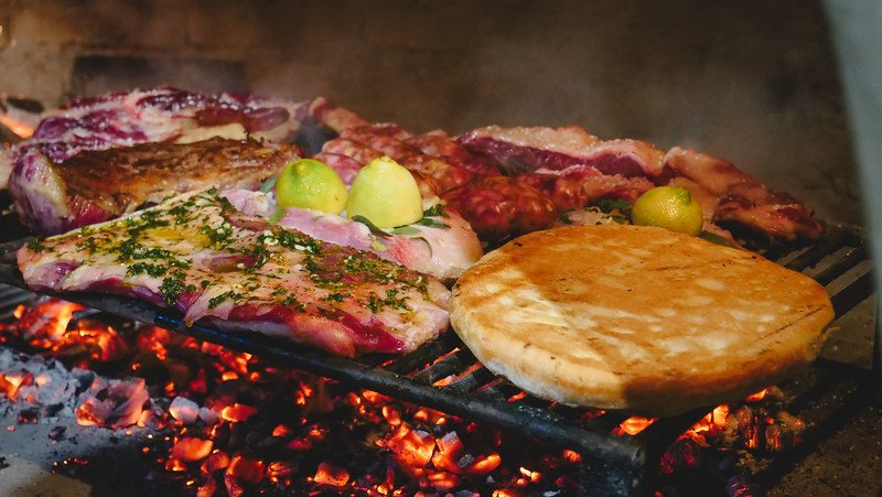 A classic Argentine asado mixed grill cooking over hot embers at Finca Adalgisa in Mendoza, Argentina. Cuts of seasoned meat, lemons, and bread are placed on the parrilla, showcasing the rich barbecue tradition that’s central to Argentine cuisine