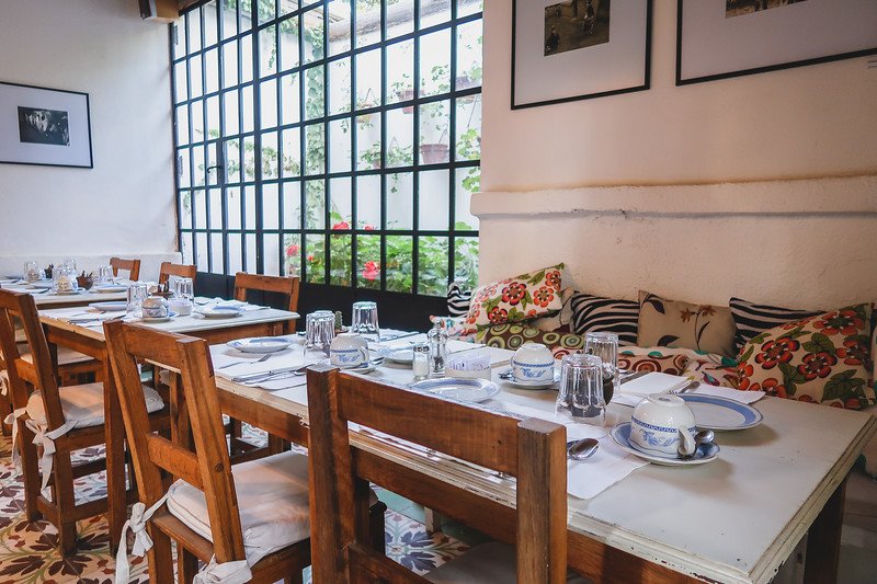 The cozy breakfast room at Finca Adalgisa in Mendoza, Argentina, set with wooden tables, floral cushions, and vintage tableware. Large grid windows let in soft morning light, creating a charming and welcoming atmosphere for guests