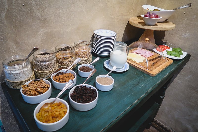 A breakfast spread at Finca Adalgisa in Mendoza, Argentina, featuring a rustic wooden table with cereals, dried fruits, nuts, seeds, milk, cold cuts, cheese, tomatoes, kiwi, and grapes. A cozy and inviting start to the day at this boutique wine hotel