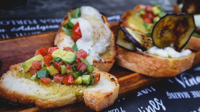 A wooden board with a selection of freshly made bruschetta served as an appetizer at Finca Adalgisa in Mendoza, Argentina. Toppings include diced avocado and tomato, mozzarella with cherry tomatoes, and grilled eggplant with creamy cheese