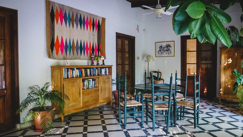 The communal area for guests at Finca Adalgisa in Mendoza, Argentina, featuring a checkered tile floor, a wooden bookshelf with colorful textiles above, and a green table with chairs. This warm and inviting space is part of the wine hotel’s interior tour