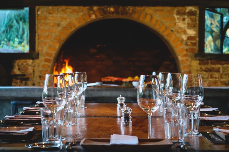 A beautifully set dinner table at Finca Adalgisa in Mendoza, Argentina, with wine glasses and cutlery neatly arranged. In the background, a roaring fire glows inside a brick oven, creating a warm and inviting atmosphere for an evening meal