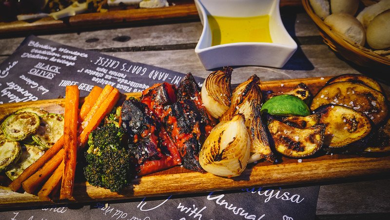 A wooden board filled with grilled vegetable tapas at Finca Adalgisa in Mendoza, Argentina. The assortment includes charred onions, carrots, zucchini, broccoli, peppers, and eggplant, served with olive oil and bread on a rustic outdoor table