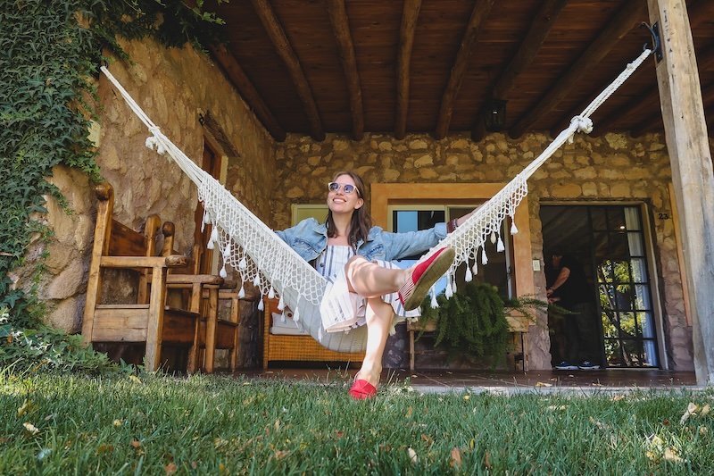 Audrey Bergner of That Backpacker relaxing on a white hammock in front of a rustic stone building at Finca Adalgisa in Mendoza, Argentina. She’s smiling, wearing a denim jacket and red espadrilles, enjoying a sunny afternoon at the boutique wine hotel