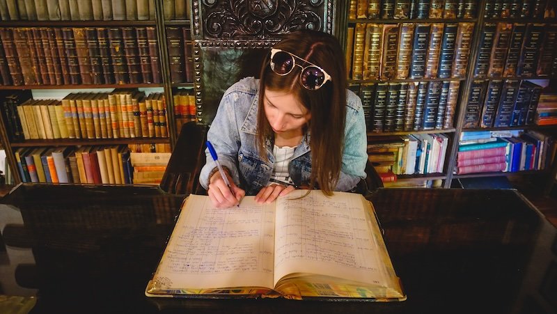 Audrey Bergner of That Backpacker signing the guestbook at Finca Adalgisa in Mendoza, Argentina. Seated at an antique wooden desk surrounded by shelves filled with old books, she captures a timeless travel moment in the hotel’s historic library