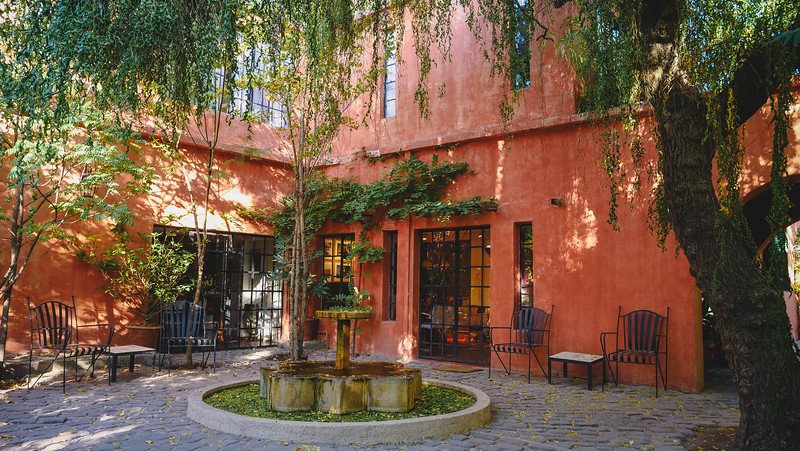 The tranquil courtyard at Finca Adalgisa, a historic wine hotel in Mendoza, Argentina, featuring a central stone fountain surrounded by wrought-iron chairs, leafy trees, and the hotel’s terracotta façade bathed in soft morning light