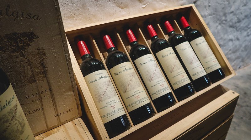 A wooden crate filled with bottles of Finca Adalgisa Malbec wine produced in Mendoza, Argentina. With a limited production of just 5,000 bottles per year, this boutique winery showcases its elegant labels and distinctive red caps in a cellar setting
