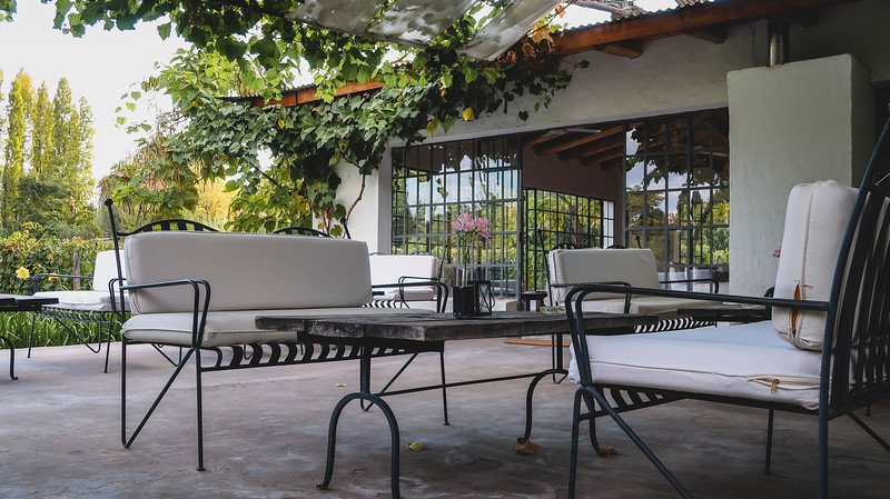 The outdoor terrace at Finca Adalgisa in Mendoza, Argentina, set for a relaxing afternoon wine o’clock. White cushioned wrought-iron chairs surround rustic tables under a vine-covered pergola, with garden views and soft natural light