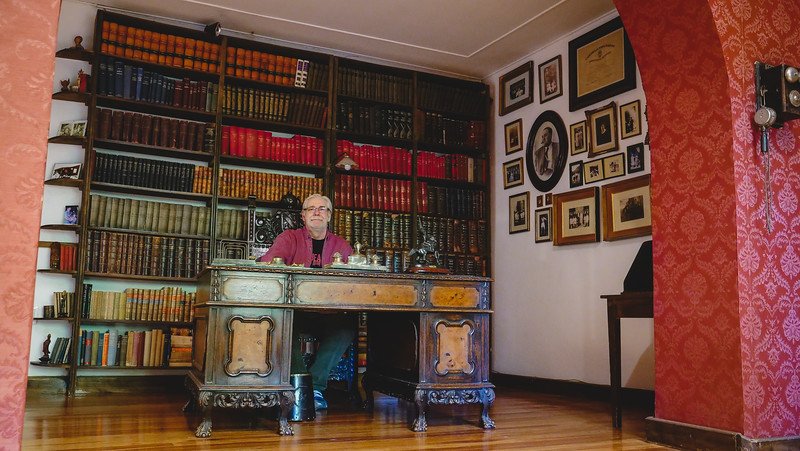 Daniel Bergner sitting at an antique wooden desk in the reading room at Finca Adalgisa in Mendoza, Argentina. The room features floor-to-ceiling bookshelves, vintage photographs, and red damask wallpaper, creating a warm and historic ambiance