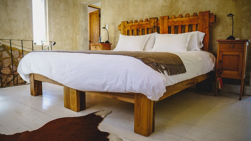 A suite room at Finca Adalgisa, a boutique wine hotel in Mendoza, Argentina, featuring a rustic wooden bed frame with crisp white linens, cozy throw blanket, and warm earth-toned walls. The minimalist decor creates a serene and inviting retreat
