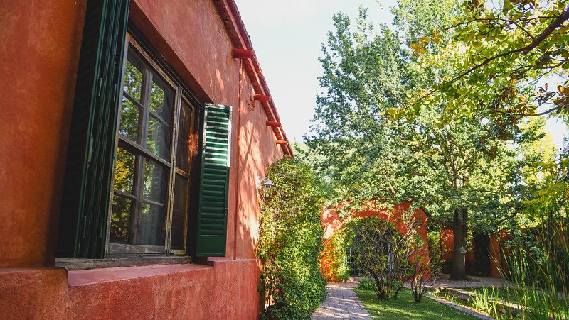 A charming window with green shutters at Finca Adalgisa in Mendoza, Argentina, overlooking the vineyard and lush garden. The terracotta walls, leafy trees, and morning sunlight create a serene and picturesque atmosphere at this boutique wine hotel