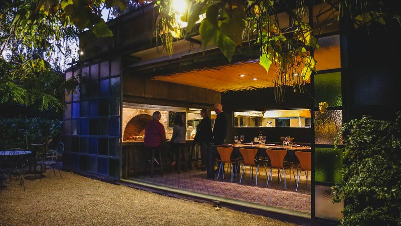 Dinner time at the winery and lounge at Finca Adalgisa in Mendoza, Argentina. Guests gather around the brick fireplace and communal tables inside a cozy, glass-walled space surrounded by greenery, enjoying an intimate evening meal with wine