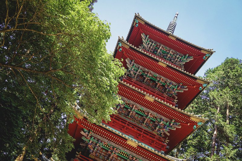 Vibrant red five-story Pagoda rising above the trees at Toshogu Shrine in Nikko, Japan, with ornate wooden carvings, gold accents, and intricate painted details shining under a clear blue sky