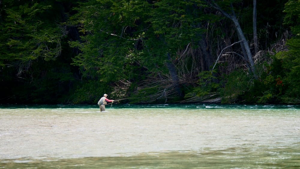 A solitary fly fisherman wades through the clear turquoise currents of a glacial river in El Bolsón, Río Negro, Argentina. Poised over a light gravel bed, the angler casts a line toward a dense green forest lining the riverbank.