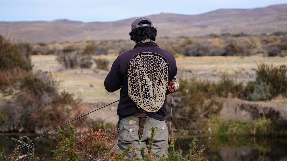 An angler seen from behind stands beside a river at Estancia Tecka, Chubut, Argentina, holding a fly rod with a visible orange reel. A woven landing net rests on his back against a backdrop of vast Patagonian scrubland and distant, arid mountains.