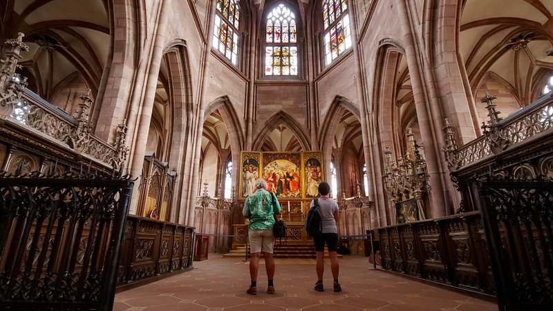 Two visitors admiring the ornate gilded altarpiece, stained glass windows, and soaring ribbed vaults inside the Gothic Freiburg Cathedral (Freiburger Münster) in Freiburg im Breisgau, Germany