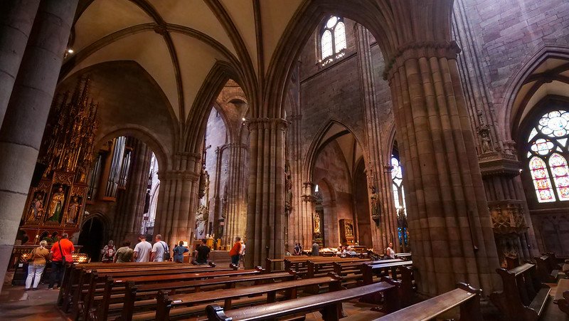 Visitors exploring the soaring vaulted arches, intricate stone columns, and stained glass windows inside the majestic Freiburg Cathedral in Freiburg im Breisgau, Germany, highlighting its impressive Gothic architecture.