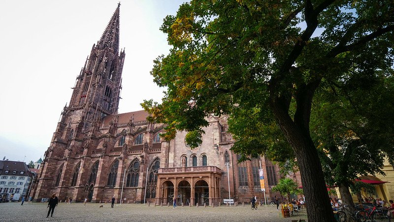 Exterior view of the towering Gothic Freiburg Cathedral (Freiburger Münster) as seen from the bustling central square in Freiburg im Breisgau, Germany, with its intricate stone spire framed by leafy trees