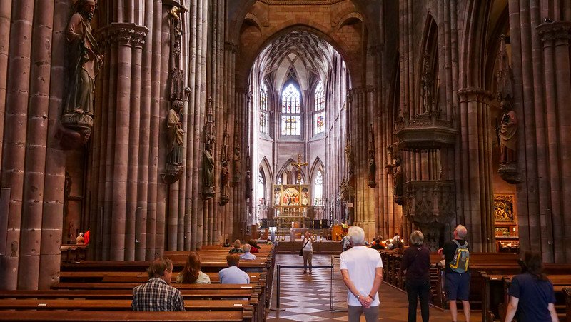 Visitors seated on wooden pews inside the grand Gothic-style Freiburg Cathedral in Freiburg im Breisgau, Germany, admiring the soaring stone columns, vaulted ceilings, and ornate altar illuminated by stained glass windows