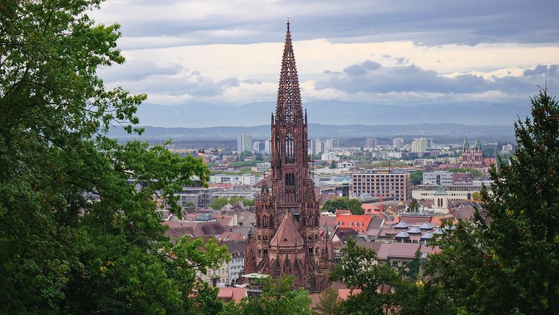Panoramic view of Freiburg im Breisgau, Germany, with the striking Gothic spire of Freiburg Cathedral rising above the city skyline, as seen from the wooded slopes of Schlossberg hill