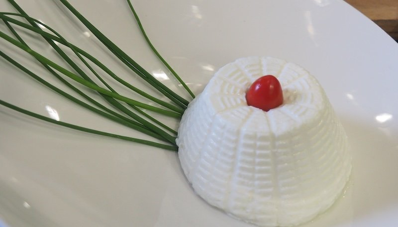 Fresh ricotta cheese molded into a delicate dome and topped with a cherry tomato, garnished with chives on a white plate, representing a simple yet elegant Italian dessert that reflects Emilia-Romagna’s artisanal dairy traditions and cuisine