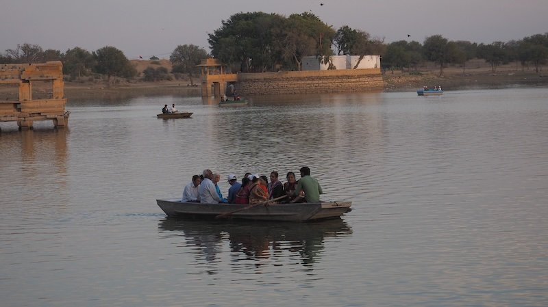 A small wooden rowboat carrying a group of passengers glides across Gadisar Lake at dusk in Jaisalmer, Rajasthan, India, with ancient sandstone pavilions and calm waters in the background