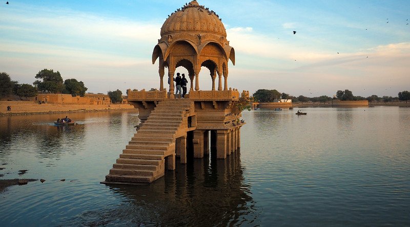 Silhouetted visitors standing inside an ornate sandstone pavilion at Gadisar Lake during sunset in Jaisalmer, India, with calm waters reflecting the warm evening light