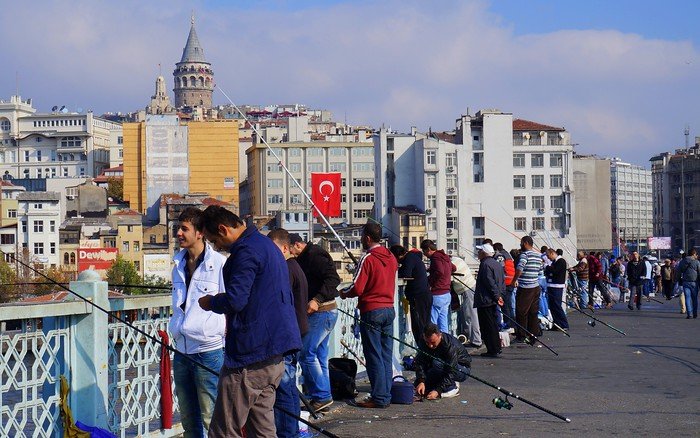 Local fishermen lined up along Galata Bridge in Istanbul, Turkey, casting their lines with the historic Galata Tower rising in the background and a Turkish flag fluttering among the city buildings