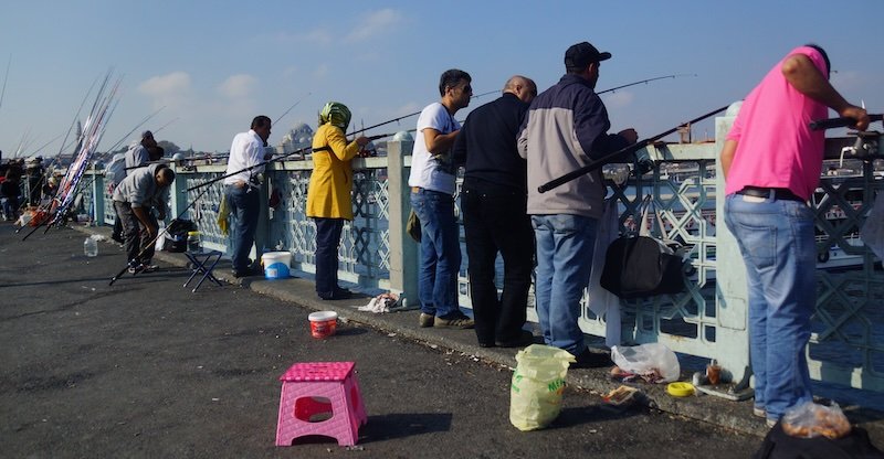 Local anglers lined up along Galata Bridge in Istanbul, Turkey, patiently fishing over the water on a sunny day, with fishing gear, stools, and tackle scattered along the walkway