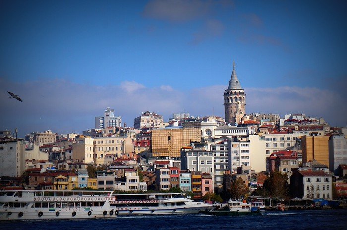 View of Galata Tower in Istanbul, Turkey, rising above a mix of colorful historic and modern buildings, with boats crossing the Golden Horn in the foreground under a clear blue sky, showcasing one of the city’s most iconic landmarks