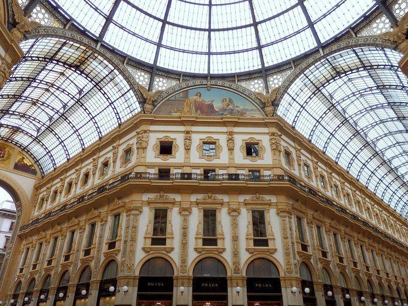 Looking up at the grand glass-and-iron dome and ornate 19th-century architecture of the Galleria Vittorio Emanuele II in Milan, Italy, showcasing intricate frescoes and luxury storefronts like Prada