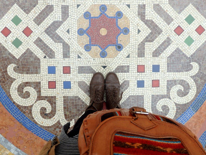 Looking down at the ornate mosaic tile floor of the Galleria Vittorio Emanuele II in Milan, Italy, with Audrey Bergner’s brown boots and backpack adding a personal travel touch