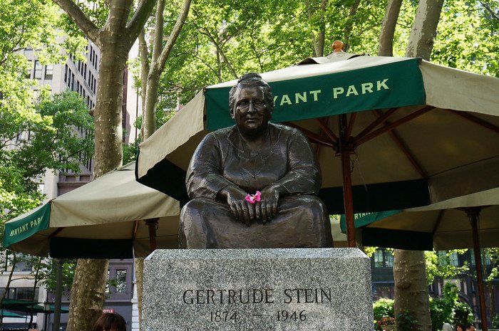 Bronze statue of Gertrude Stein seated with folded hands, holding a small pink flower, at Bryant Park in New York City. The statue is set against green trees and iconic Bryant Park umbrellas, located behind the New York Public Library on a sunny summer day