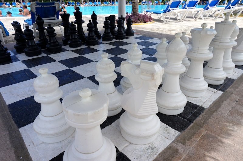 Oversized black and white chess pieces arranged on a giant outdoor chessboard beside a sparkling resort pool at Cayo Santa María, Cuba, offering a fun leisure activity during a tropical honeymoon escape