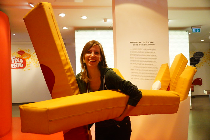 Giant french fries inside the Currywurst Museum in Berlin, Germany as being held by a laughing and smiling That Backpacker Audrey Bergner