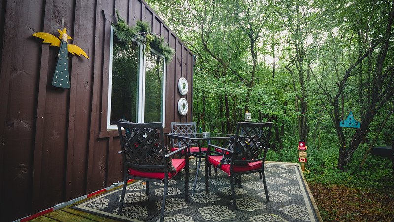 The outdoor patio of a cosy tiny cabin Airbnb in Glen Huron, Ontario, Canada, surrounded by lush green forest. The patio features a patterned outdoor rug, a glass table with four black chairs with red cushions, whimsical decorations, and peaceful woodland views