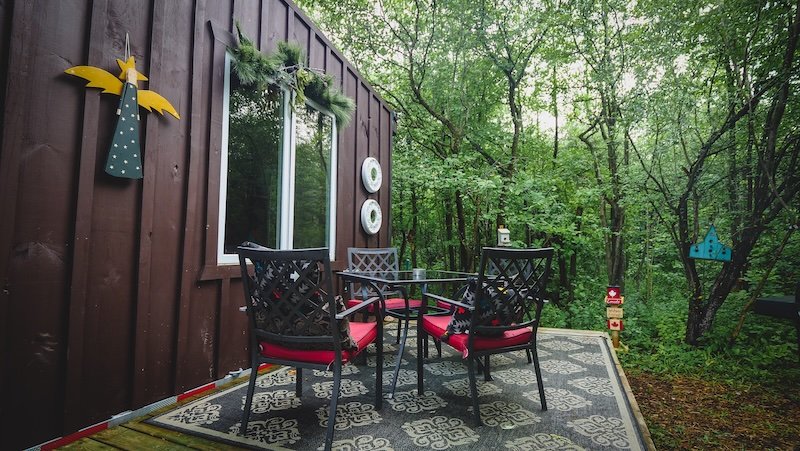 The charming outdoor patio of a tiny cabin in Glen Huron, Ontario, surrounded by lush forest. A cozy seating area with black chairs and red cushions sits on a patterned rug, creating the perfect spot to relax and enjoy nature during a woodland getaway