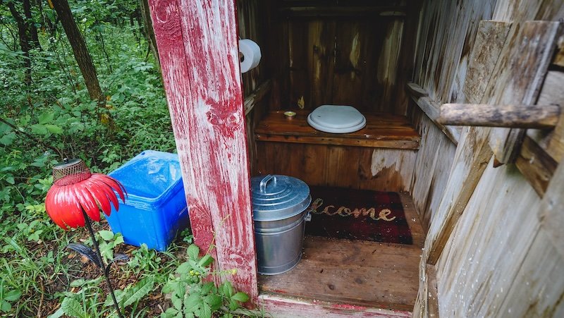 Inside view of a rustic wooden outhouse at a tiny cabin getaway in Glen Huron, Ontario, Canada. The space features a simple toilet seat on a wooden bench, a welcome mat, a small trash bin, and a toilet paper roll, blending outdoor charm with basic comfort