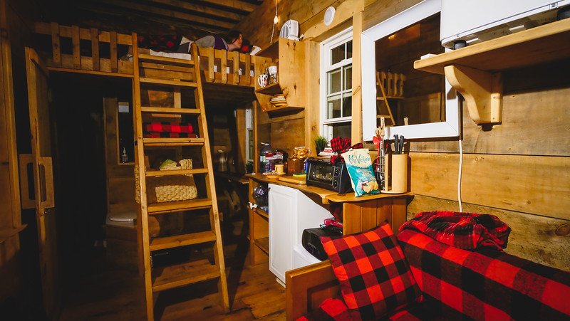 A warm and inviting interior of a tiny cabin in Glen Huron, Ontario, featuring wood panel walls, a cozy loft bedroom accessed by a ladder, a compact kitchen, and red-and-black plaid accents on the sofa and bedding that enhance the rustic charm