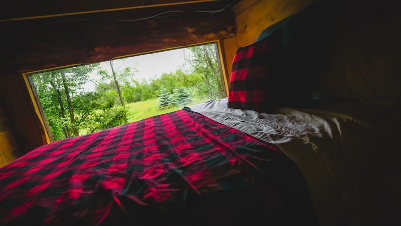 The cosy loft bed inside a tiny cabin Airbnb in Glen Huron, Ontario, Canada, features a red-and-black plaid blanket and a large picture window overlooking a peaceful forest and meadow, creating a serene and rustic atmosphere perfect for a nature getaway