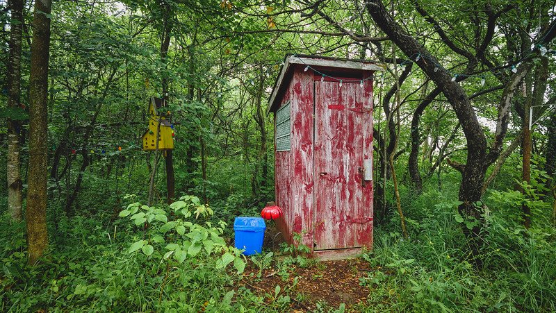 Rustic red wooden outhouse surrounded by lush green forest at a tiny cabin getaway in Glen Huron, Ontario, Canada. The charmingly weathered structure stands beside a blue water container, blending into the peaceful woodland retreat setting