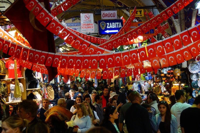 Crowds of shoppers exploring the vibrant stalls of the Grand Bazaar in Istanbul, Turkey, under colorful Turkish flags and surrounded by textiles, lamps, and souvenirs