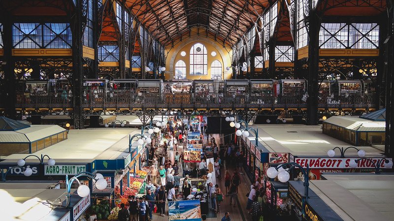 The bustling interior of the Great Market Hall in Budapest, Hungary, filled with colorful produce stands, local food vendors, and souvenir stalls under a striking iron and glass roof. Visitors browse the lively market, a must-see attraction in the city
