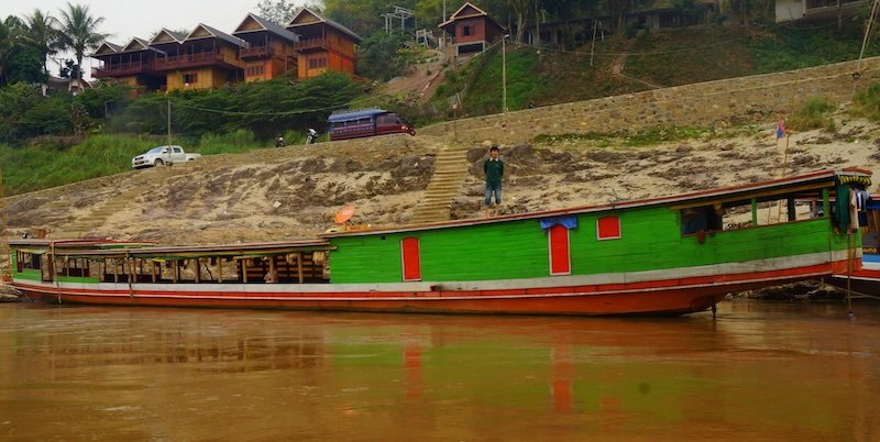 A vibrant green and red slow boat is docked along the sandy banks of the Mekong River in Laos, with wooden guesthouses and a steep staircase leading down to the water in the background. This classic vessel is used for the scenic two-day journey to Luang Prabang