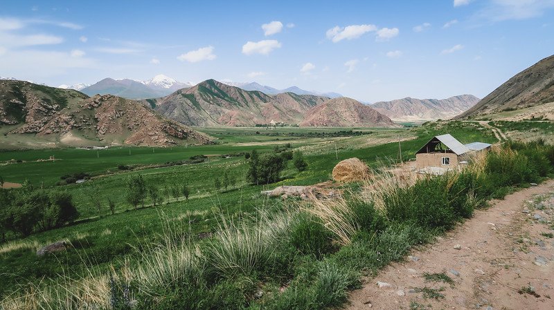 A scenic green valley surrounded by rugged mountains with a small farmhouse and haystack along a dirt path on the way back to town during the 3-Day Panorama Trek in Kyrgyzstan