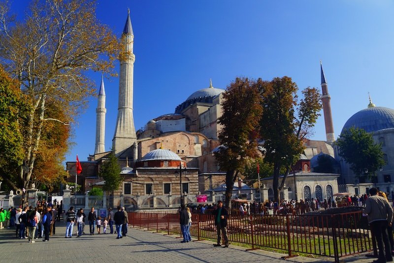 Visitors strolling through the courtyard of the majestic Hagia Sophia in Istanbul, Turkey, with its towering minarets and grand domes framed by autumn trees under a clear blue sky