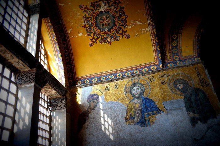 Interior mosaics of Hagia Sophia in Istanbul, Turkey, depicting religious figures with gold backgrounds, illuminated by sunlight streaming through arched windows, showcasing the historic Byzantine artwork and spiritual atmosphere of the monument